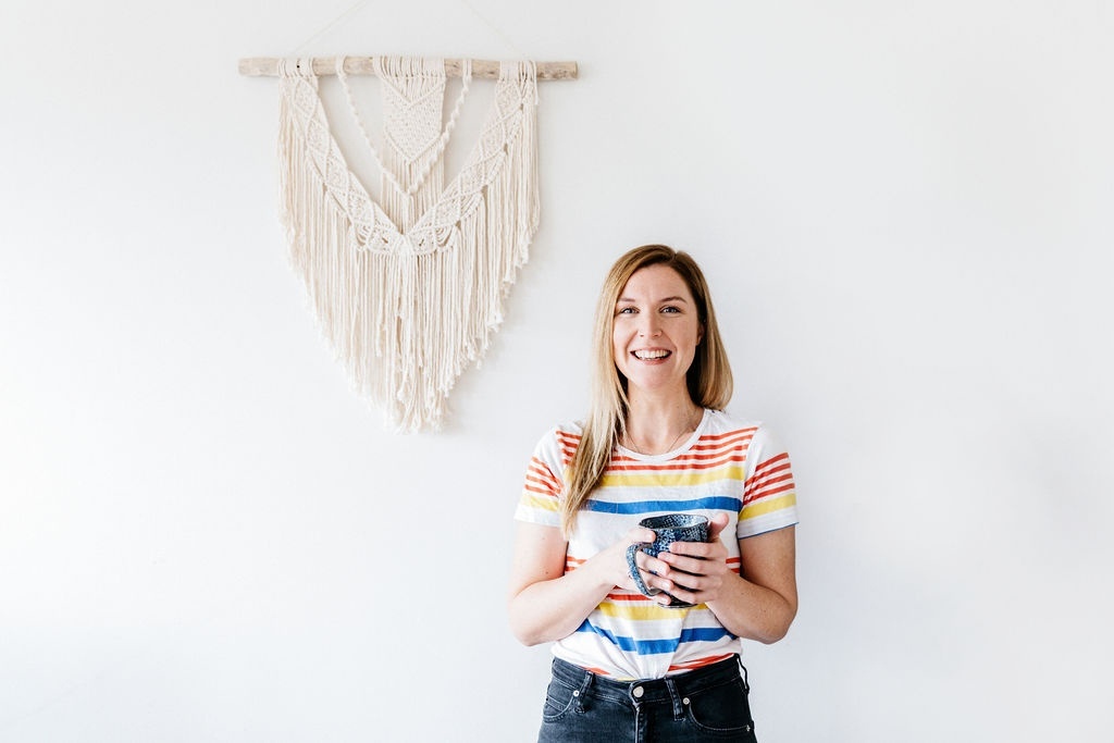 Natalie is laughing while holding a cup of coffee. She is wearing a white t-shirt ith red, yeelow and blue stripes and black jeans. In the background is a Macrame hanging on a white wall.