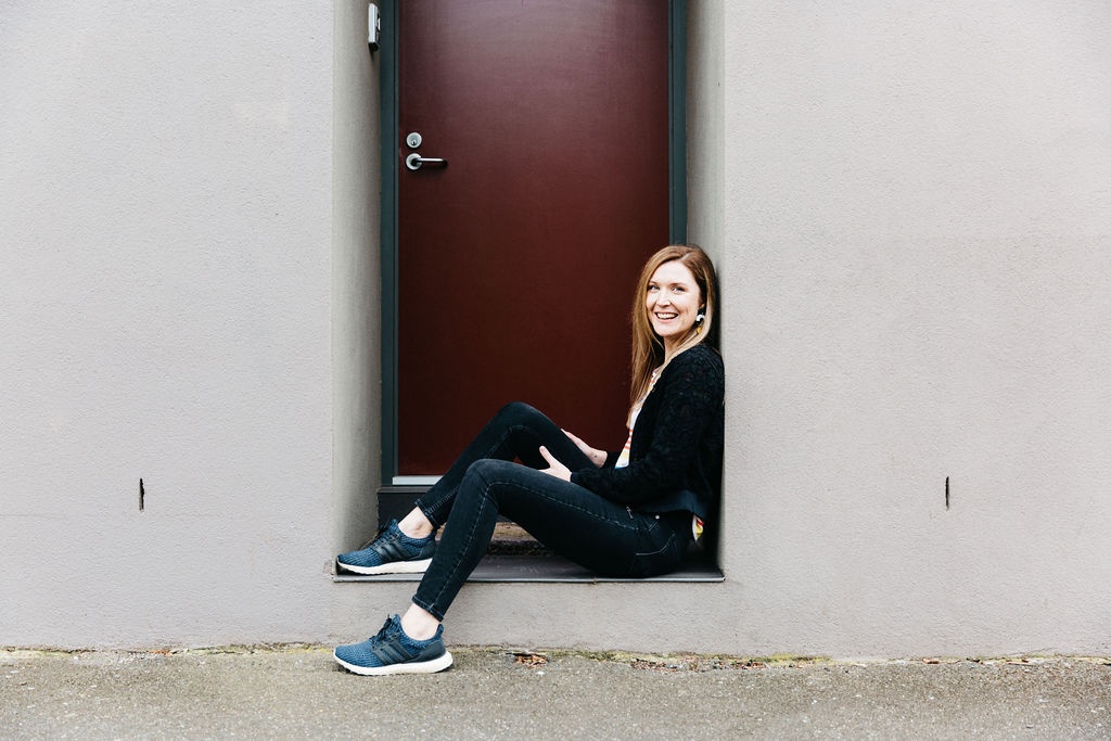 Natalie is sat in a concreate grey doorway, in front of a marron red door, she is wearing black jeans and a black jacket with blue adidas parley running shoes. These are the the 5 Lessons I Wish I Learned Before I Started My Business.